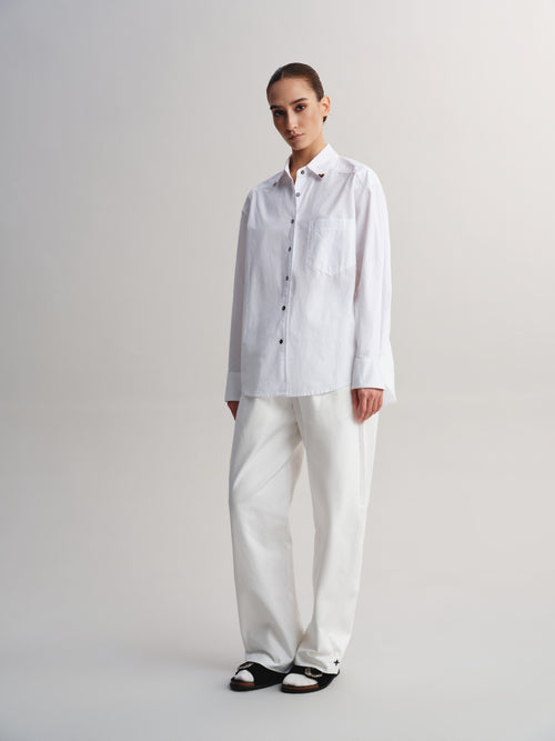 A model stands against a light gray studio background wearing a white long-sleeve button-up shirt with a chest pocket, white wide-leg trousers, and black slide sandals.