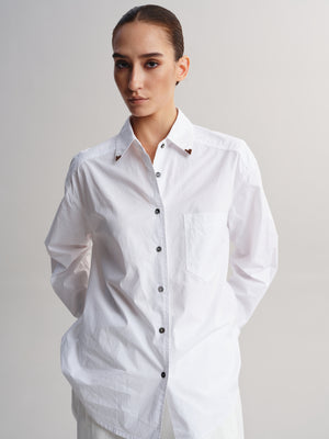 A woman modeling a white long-sleeve button-up shirt with a chest pocket and small brown decorative accents near the collar, photographed against a plain light gray studio background.