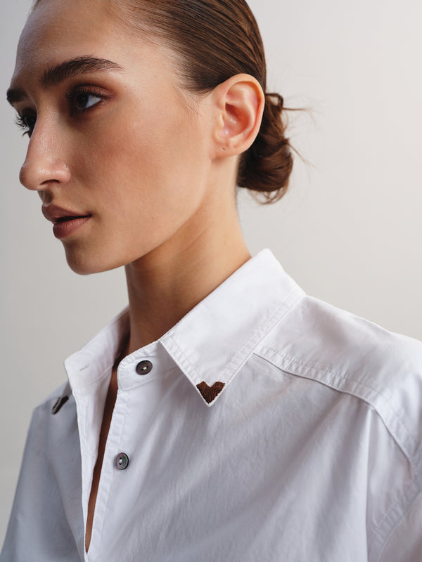 Close-up of a woman wearing a white button-up shirt with a pointed collar and a small brown heart-shaped chest emblem, photographed against a plain light background.