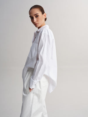 A model wearing a white long-sleeve button-up shirt with a chest pocket and white trousers, photographed against a neutral light gray studio background.