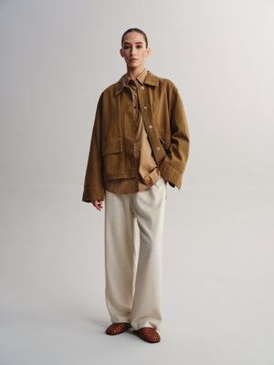 A model stands against a light gray studio background wearing a brown corduroy utility jacket over a beige shirt, paired with wide-leg cream trousers and brown woven shoes.