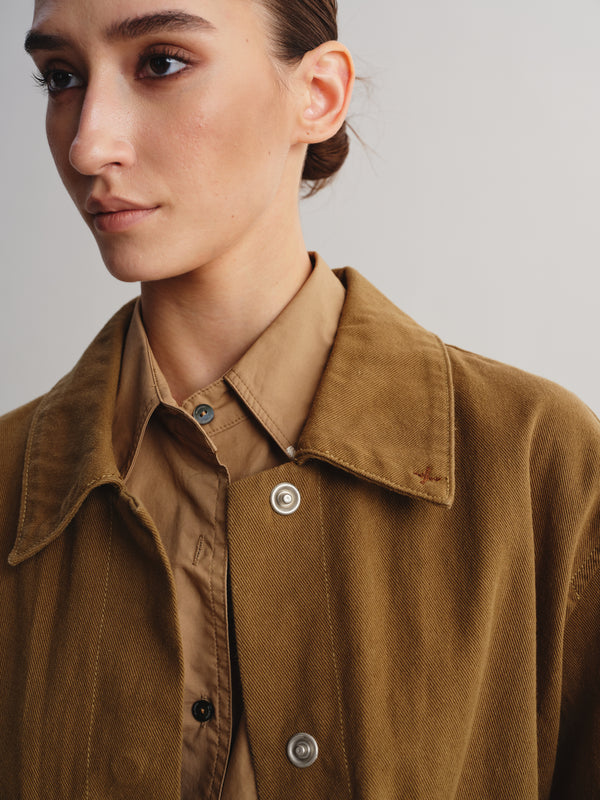 Close-up of a model wearing a brown suede-like button-up shirt jacket over a tan collared shirt, against a plain light gray background.