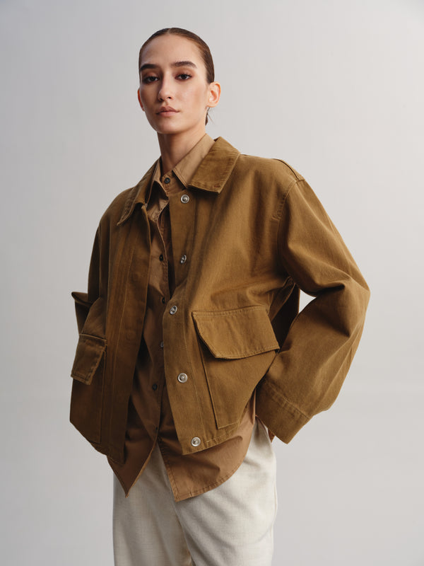 A model wearing a brown corduroy utility jacket over a matching brown shirt and off-white trousers, photographed against a neutral light gray studio background.