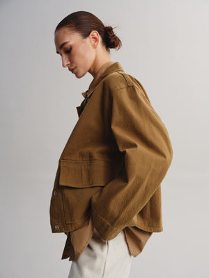 A model wearing a brown utility jacket with a relaxed fit, paired with off-white trousers, photographed against a neutral light gray background.