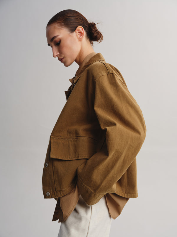 A model wearing a brown utility jacket with a relaxed fit, paired with off-white trousers, photographed against a neutral light gray background.