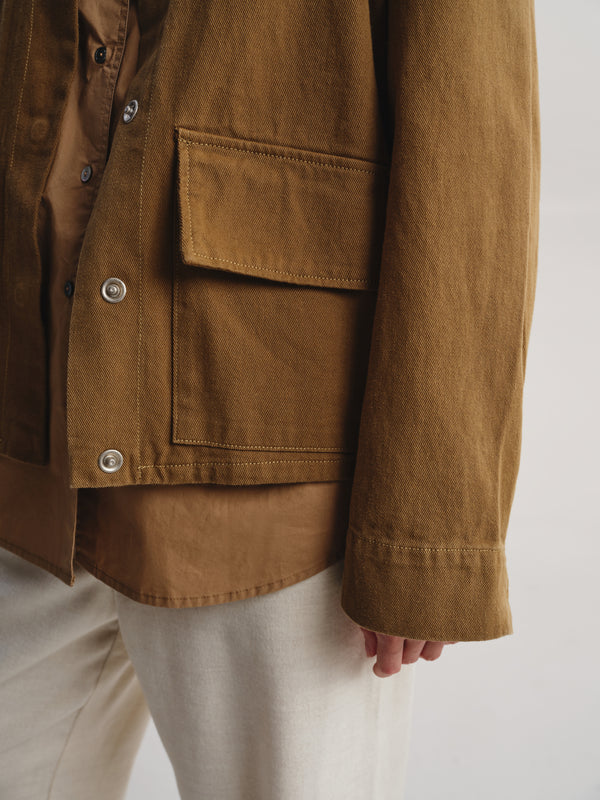 Close-up of a person wearing a brown jacket over a brown shirt and off-white pants, with the jacket's chest pocket and buttons visible against a plain light background.