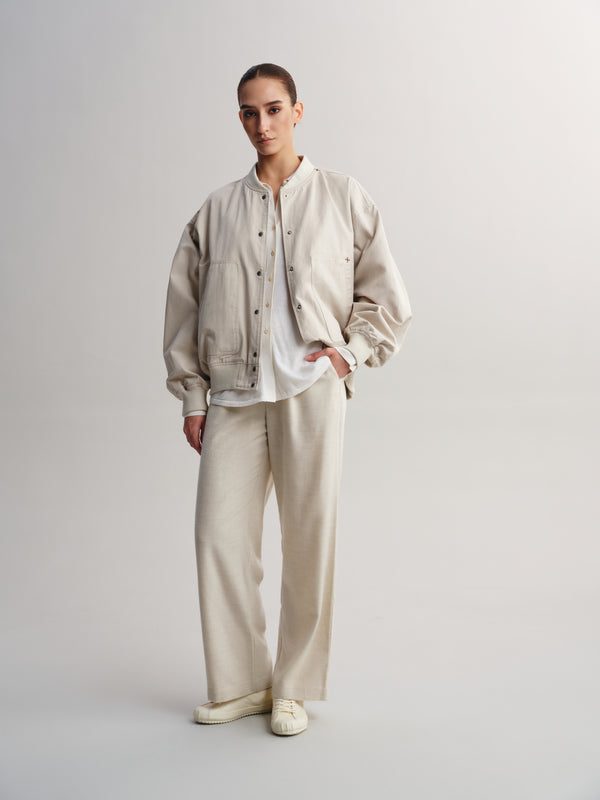 A model stands against a light gray studio background wearing a light beige oversized button-front jacket over a white shirt, matching beige wide-leg trousers, and off-white slip-on shoes.