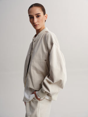 A model wearing a light beige oversized bomber jacket with a front zipper and ribbed collar, cuffs, and hem, paired with matching beige trousers, set against a plain light gray studio background.