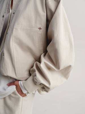 Close-up of a person wearing a light beige overshirt with a small embroidered cross detail on the chest pocket, paired with a white shirt and matching beige trousers against a plain light background.