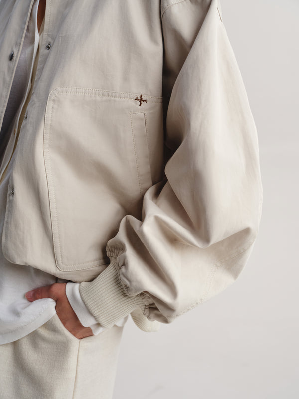 Close-up of a person wearing a light beige overshirt with a small embroidered cross detail on the chest pocket, paired with a white shirt and matching beige trousers against a plain light background.