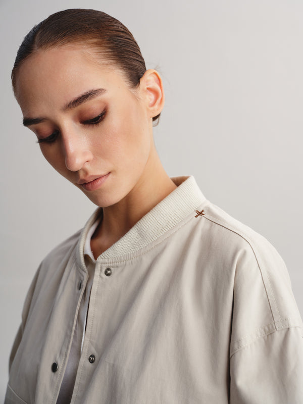 A close-up shot of a model wearing a light beige button-up shirt with a ribbed collar and a small embroidered star detail on the shoulder, set against a plain light gray background.