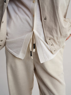 A close-up of a person wearing a light beige overshirt over a white shirt and beige trousers, with the drawstring of the shirt visible.