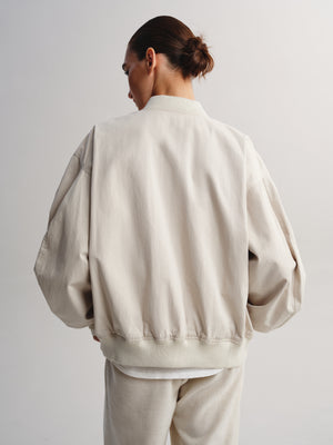 A model shown from behind wearing a light beige oversized bomber jacket and matching beige trousers against a plain light gray studio background.