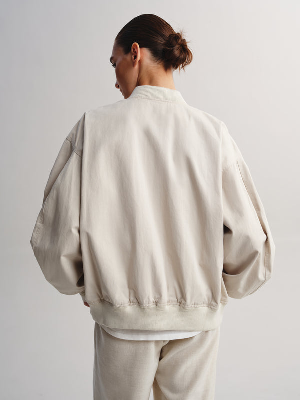 A model shown from behind wearing a light beige oversized bomber jacket and matching beige trousers against a plain light gray studio background.