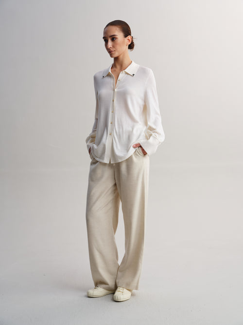 A woman modeling a white long-sleeve button-up shirt and beige wide-leg trousers against a plain light gray studio background.