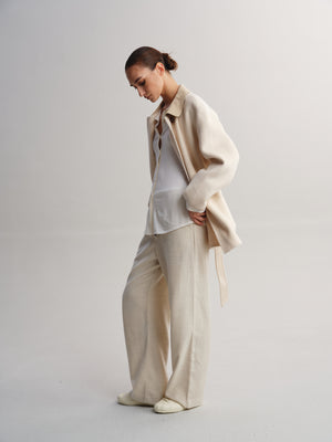 A model wearing a light beige oversized jacket over a white shirt and matching wide-leg trousers, standing against a neutral light gray studio background.