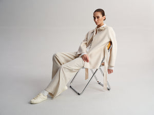 A model sits on a folding chair wearing a cream-colored tailored suit with a matching tie and white sneakers against a light gray studio background.