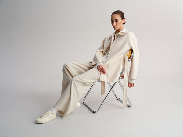 A model sits on a folding chair wearing a cream-colored tailored suit with a matching tie and white sneakers against a light gray studio background.