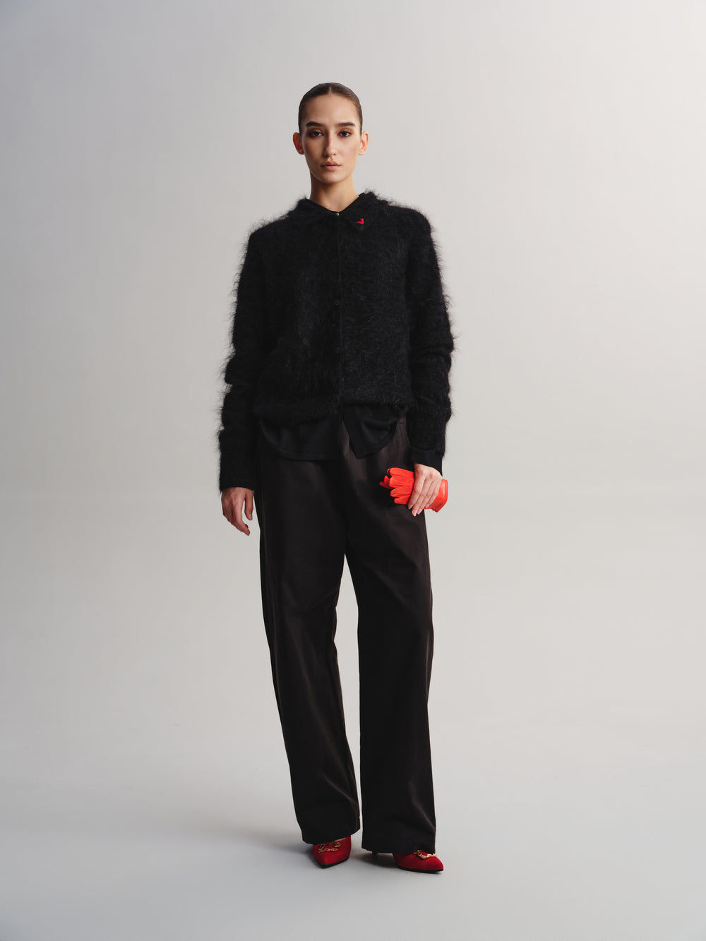 A model stands against a light gray studio background wearing a black fuzzy cropped jacket, black wide-leg trousers, and red shoes, while holding a small bright red pouch.