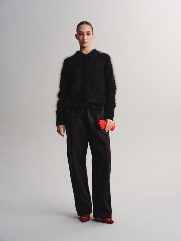 A model stands against a light gray studio background wearing a black fuzzy cropped jacket, black wide-leg trousers, and red shoes, while holding a small bright red pouch.