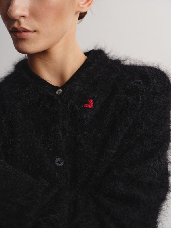 Close-up of a model wearing a black fuzzy mohair-style cardigan with a small red embroidered logo on the chest.