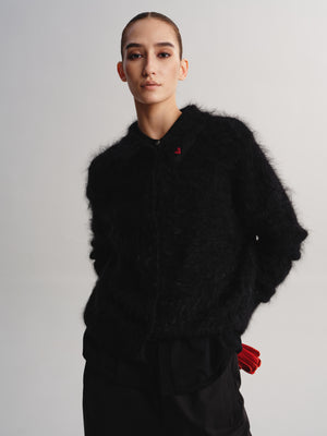 A model wearing a black fuzzy textured jacket with a small red embroidered logo on the chest, paired with black pants and red gloves, against a light gray studio background.