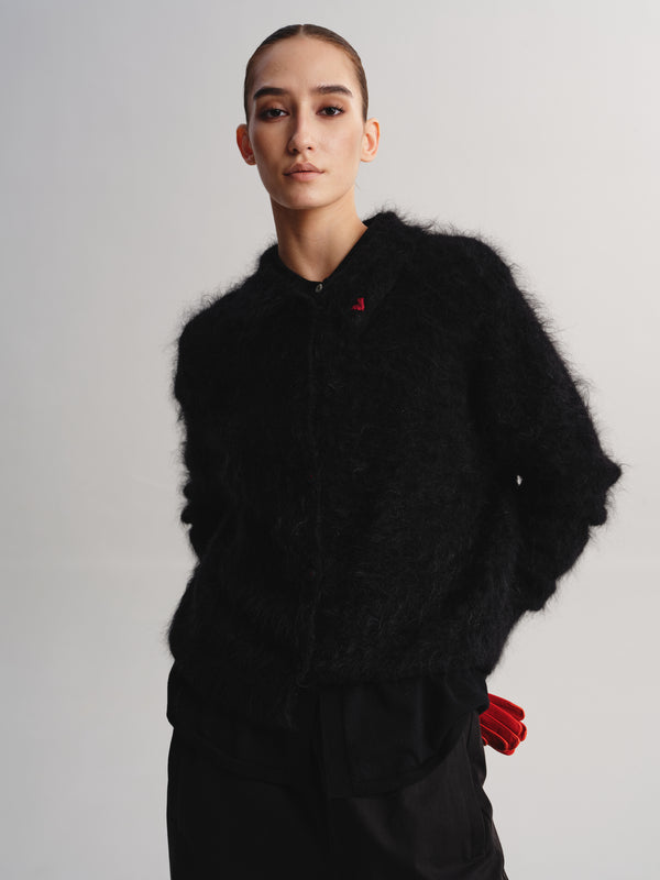 A model wearing a black fuzzy textured jacket with a small red embroidered logo on the chest, paired with black pants and red gloves, against a light gray studio background.