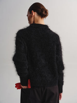A model shown from the back wearing a black fuzzy mohair-style knit sweater, paired with black trousers and holding a red clutch purse, against a plain light gray studio background.