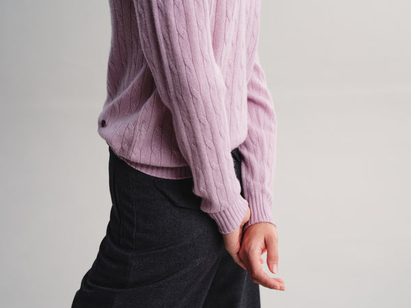A person wearing a light pink cable-knit sweater and dark charcoal gray trousers, shown from the side against a plain light gray studio background.