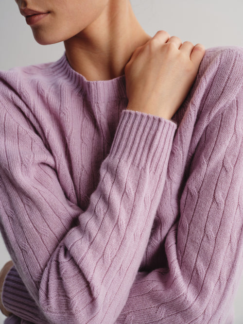 A close-up of a person wearing a dusty lavender cable-knit sweater, showing the ribbed crew neck and textured sleeves against a neutral light gray background.