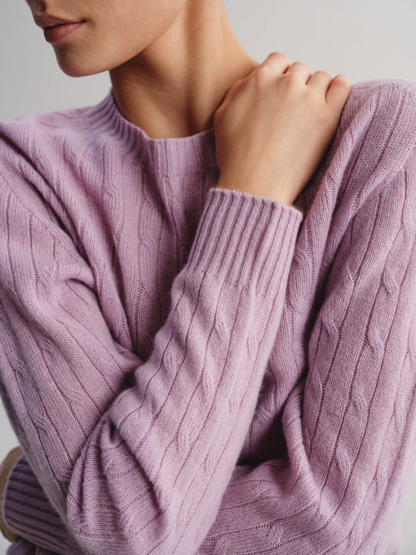 A close-up of a person wearing a dusty lavender cable-knit sweater, showing the ribbed crew neck and textured sleeves against a neutral light gray background.