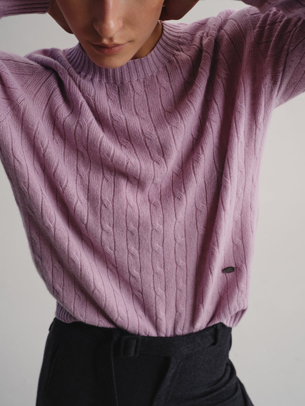 A close-up of a person wearing a dusty lavender cable-knit sweater with dark trousers, against a plain light background.