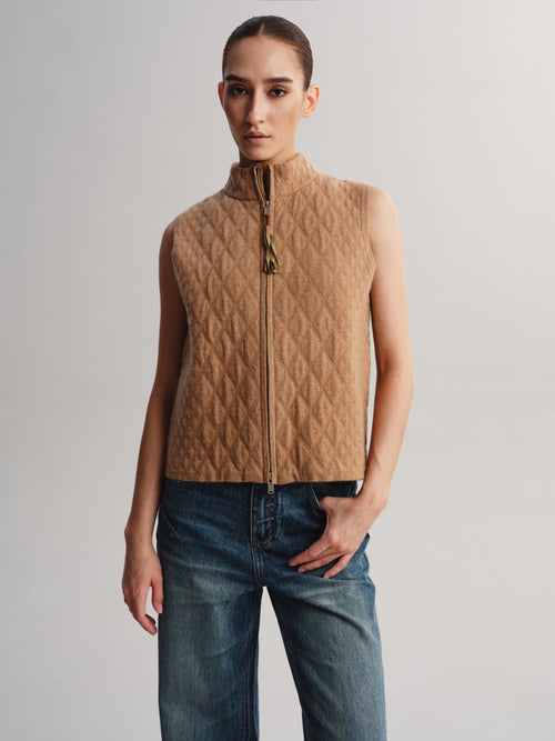A model wearing a sleeveless, quilted camel-brown zip-up vest with a high collar, paired with blue jeans, against a plain light gray studio background.