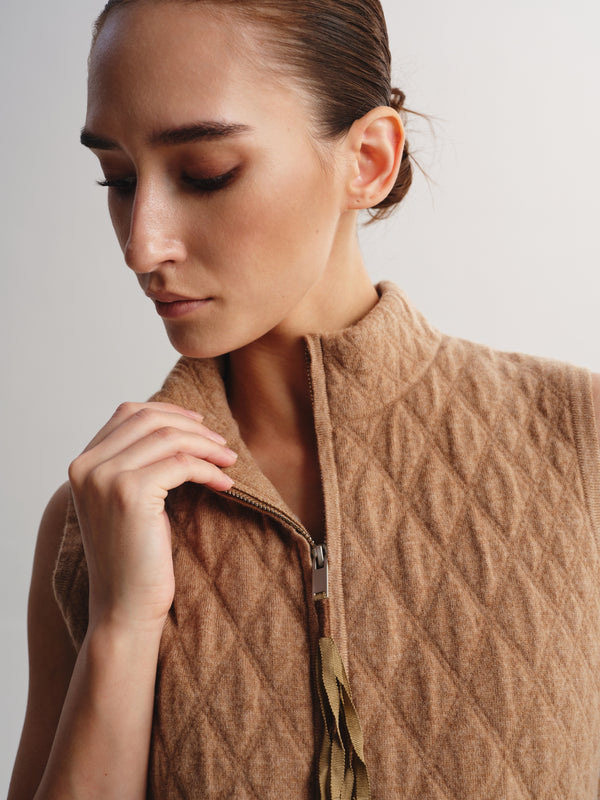 A close-up of a woman wearing a sleeveless quilted camel-brown vest with a high stand-up collar and a front zipper, photographed against a plain light background.