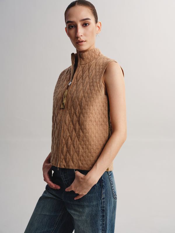 A woman modeling a sleeveless quilted zip-front vest in a warm camel brown color, paired with blue jeans, against a plain light gray studio background.