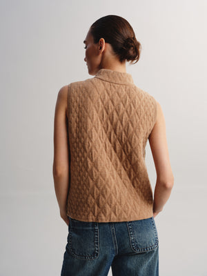 A woman shown from the back wearing a sleeveless, quilted camel-brown vest over a light top and blue jeans, against a plain light gray studio background.