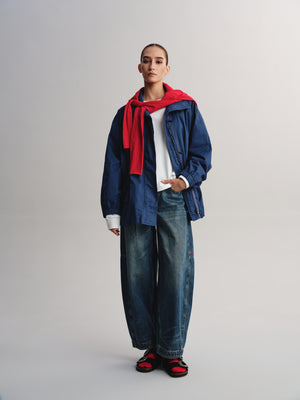 A model stands against a light gray studio background wearing an oversized blue denim jacket over a white shirt, paired with wide-leg blue jeans, a bright red scarf, and red-and-black sandals.