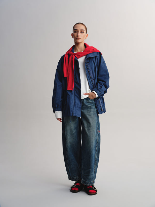 A model stands against a light gray studio background wearing an oversized blue denim jacket over a white shirt, paired with wide-leg blue jeans, a bright red scarf, and red-and-black sandals.