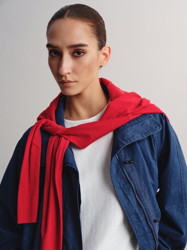 A model wearing a vibrant red knit scarf wrapped around her neck, layered over a white t-shirt and a blue denim jacket, against a plain light gray background.