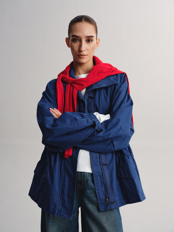 A model wearing a navy blue utility jacket with a bright red hood, layered over a white t-shirt and blue jeans, standing against a light gray studio background.
