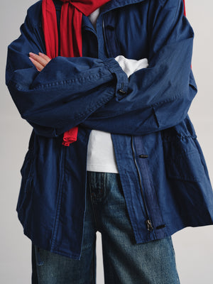 A person wearing an oversized navy blue utility jacket over a white shirt and red scarf, paired with blue jeans, shown from the chest down against a plain light gray background.