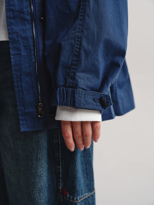 Close-up of a person wearing a dark blue lightweight jacket over a white shirt and blue jeans, with the hand and cuff of the jacket visible against a plain light background.