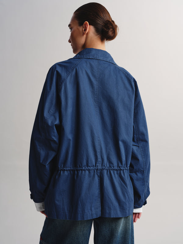 A model shown from the back wearing a blue utility jacket with a gathered waist and wide sleeves, paired with blue jeans against a light gray studio background.