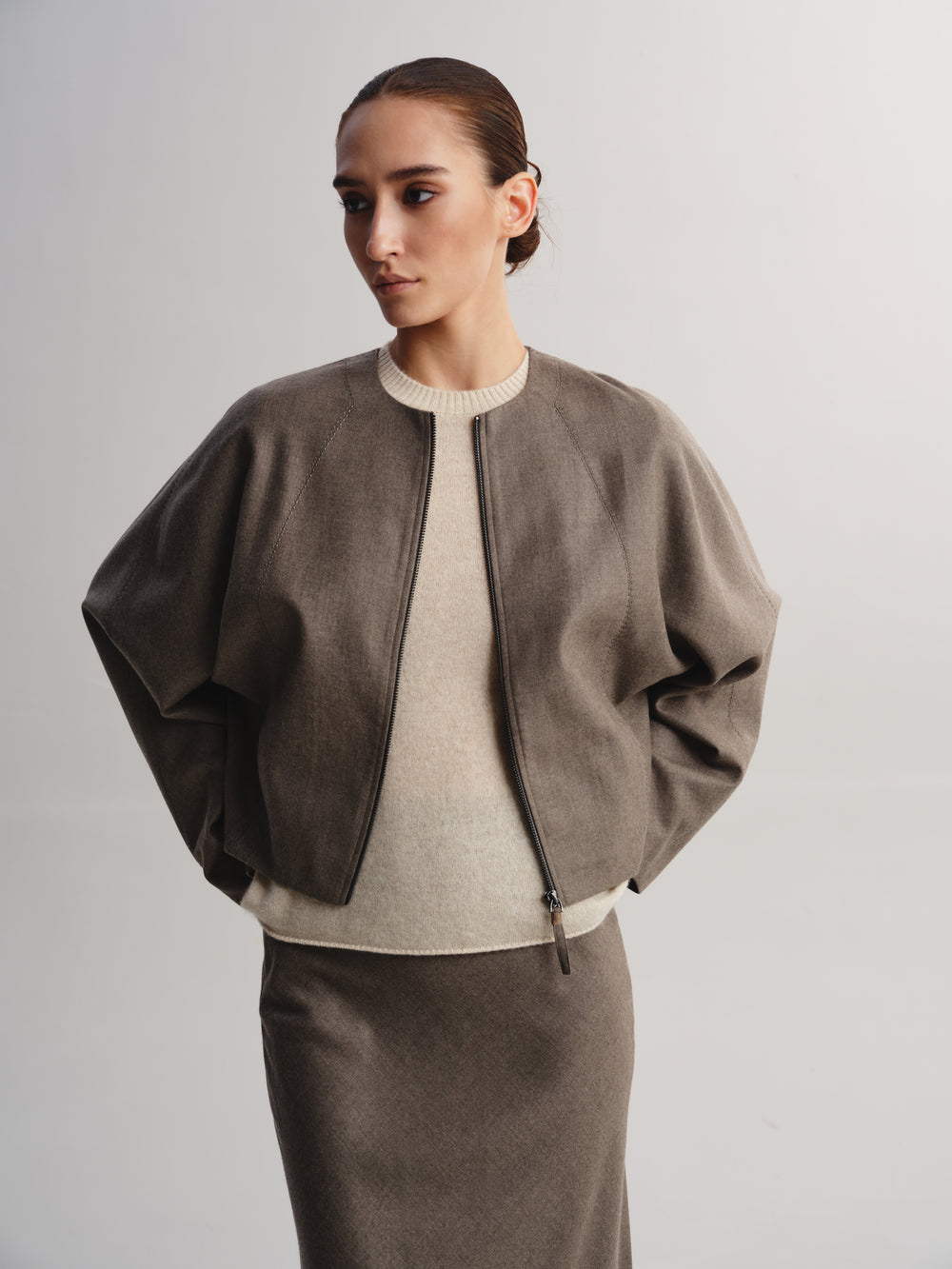 A woman modeling a taupe cropped bomber jacket over a cream ribbed sweater and a matching taupe skirt, photographed against a neutral light gray studio background.