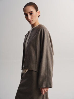 A model wearing a taupe-brown cropped jacket with wide draped sleeves and a matching skirt, photographed against a neutral light gray studio background.