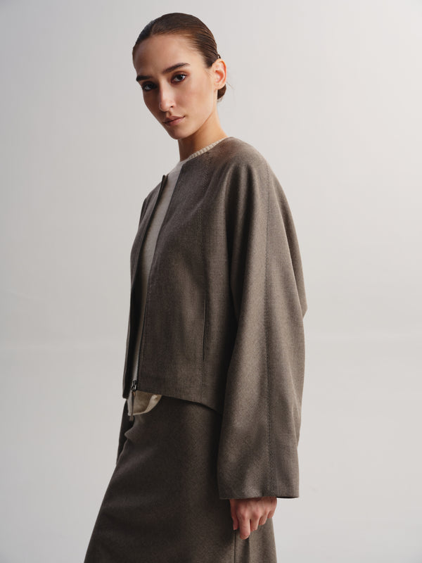 A model wearing a taupe-brown cropped jacket with wide draped sleeves and a matching skirt, photographed against a neutral light gray studio background.