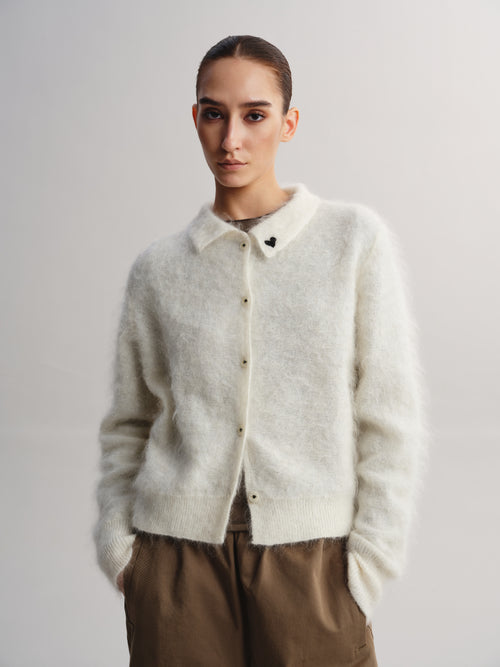A model wearing a fuzzy, off-white knit cardigan with a collar and button front, paired with brown trousers, against a plain light gray studio background.