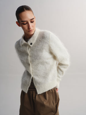 A model wearing a fuzzy, light cream-colored knit cardigan with a collar and front buttons, paired with brown trousers, against a plain light gray studio background.