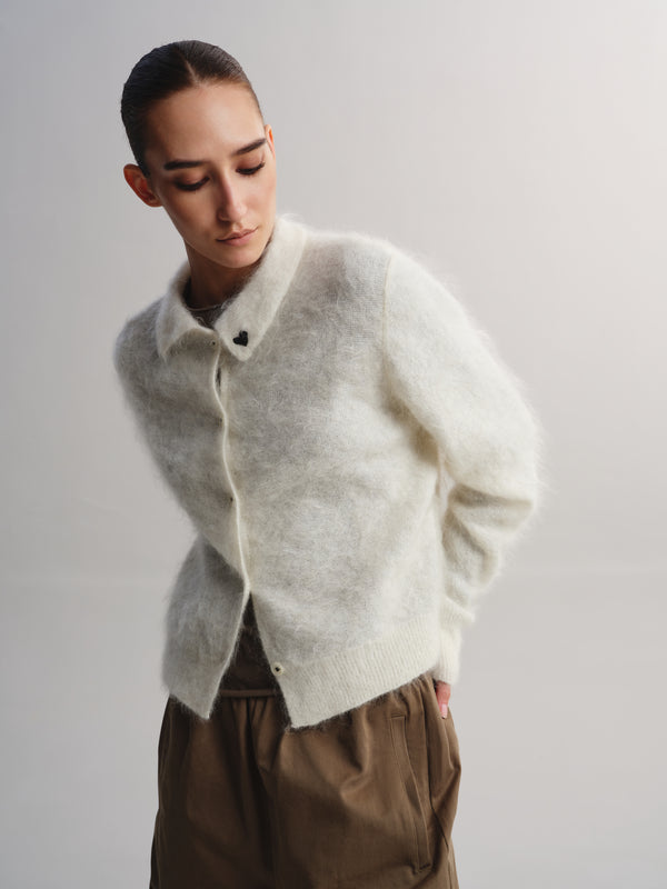 A model wearing a fuzzy, light cream-colored knit cardigan with a collar and front buttons, paired with brown trousers, against a plain light gray studio background.
