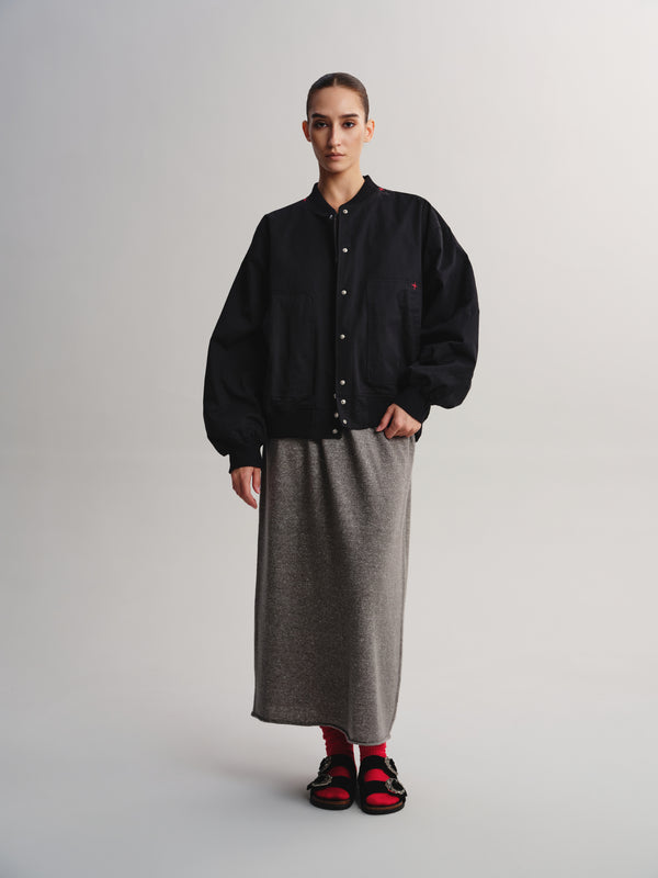 A model stands against a light gray studio background wearing a black button-front jacket with a small red embroidered logo on the chest, a long gray textured skirt, red socks, and black sandals with red straps.